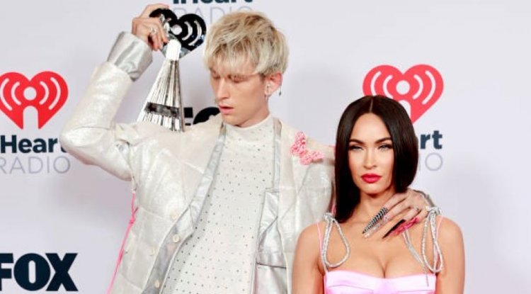 Wearing makeup, with long nails and a pink butterfly on his shoulder - MGK